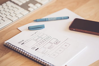A neat workspace featuring a sketchpad with wireframes, a smart, and a keyboard on a wooden desk. HD wallpaper