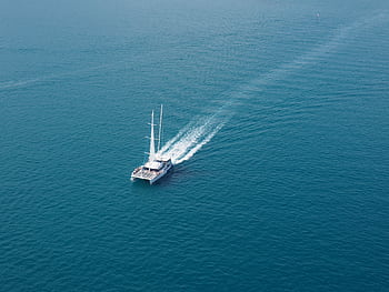 A sailboat cruises through clear blue ocean waters, captured from an aerial perspective. HD wallpaper
