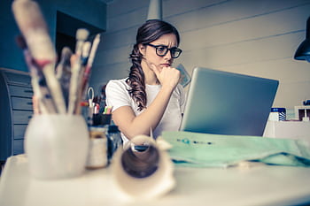 Young woman with glasses deeply focused on a laptop surrounded by art supplies in a home office. HD wallpaper