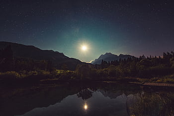 A peaceful view of moonlit mountains reflected in a tranquil lake under a starry sky. HD wallpaper