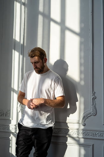 A thoughtful bearded man in a white t-shirt stands in a sunlit room with ornate walls. HD phone wallpaper