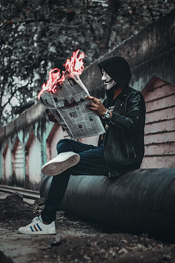 A person wearing a mask reads a flaming newspaper on an urban street, creating a surreal and mysterious scene. HD phone wallpaper
