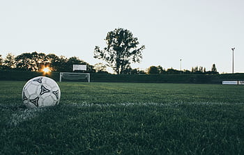 Soccer ball on field at sunrise, serene landscape with goalpost in background. HD wallpaper