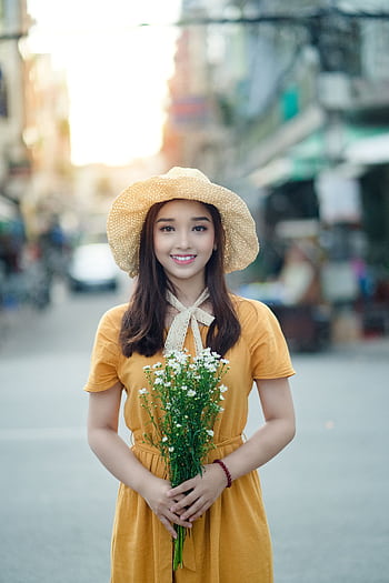 Smiling woman in a yellow dress and straw hat holding flowers on a sunlit street. HD phone wallpaper