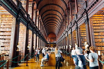 Visitors explore a grand library hallway with towering bookshelves in an iconic university. HD wallpaper
