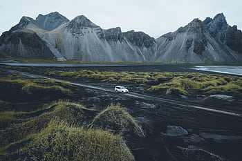 A white car traverses a gravel road against Iceland's dramatic mountain backdrop. HD wallpaper