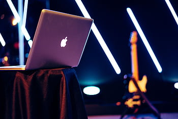 A sleek laptop sits on a table with vibrant neon lights and a guitar in the background. HD wallpaper