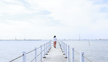 A woman stands quietly on a serene pier overlooking the calm ocean under a cloudy sky. HD wallpaper