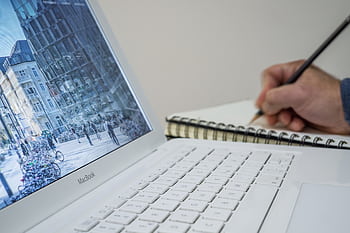 A person taking notes in a notebook beside an open laptop displaying an urban scene. HD wallpaper