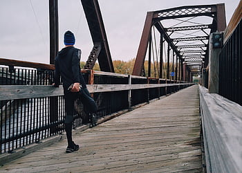 Man stretching on a wooden footbridge during a daytime run on an iron bridge, emphasizing fitness and recreation. HD wallpaper