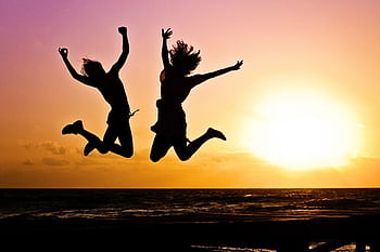 Silhouettes of two people joyfully jumping on a beach during a vibrant sunset. HD wallpaper