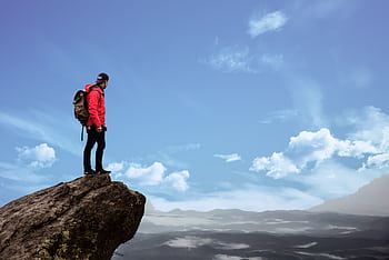 Hiker in red jacket stands on a cliff edge, overlooking a scenic mountain view under a bright blue sky. HD wallpaper