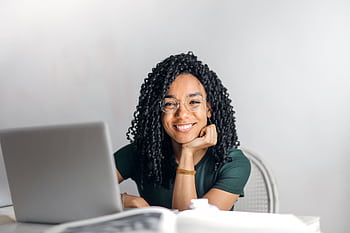 Joyful businesswoman with curly hair smiling at camera while using laptop indoors. HD wallpaper