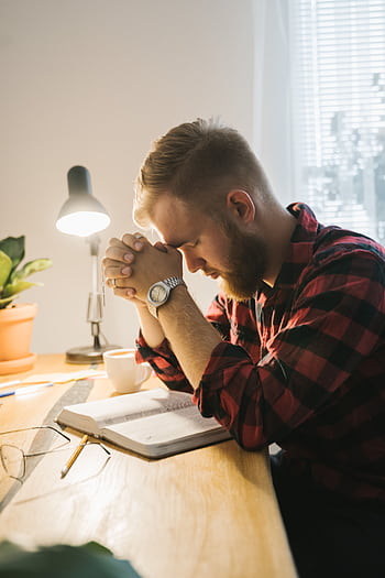 Bearded man in plaid shirt praying with closed eyes at desk with Bible under soft indoor lighting. HD phone wallpaper
