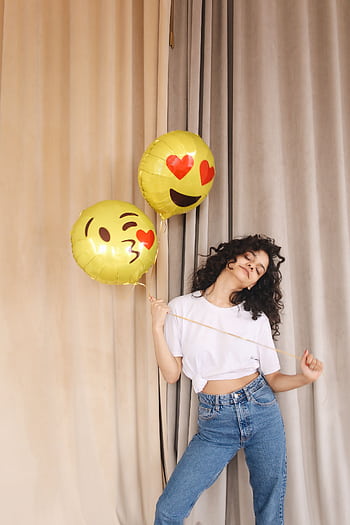 Cheerful woman in jeans and white shirt holding emoji balloons, enjoying a celebration indoors. HD phone wallpaper
