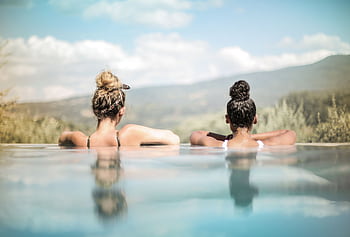 Two women relaxing in a pool with scenic mountain and countryside views. HD wallpaper