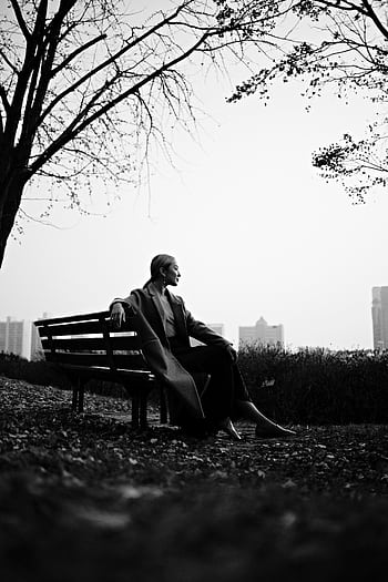 Black and white of a woman sitting on a park bench looking contemplative. Urban setting. HD phone wallpaper