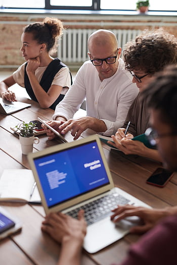 Diverse team engaged in a collaborative business meeting around a wooden table with laptops. HD phone wallpaper