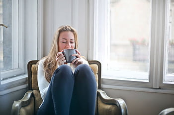 Woman enjoying a warm drink in a cozy armchair by a window, embracing relaxation. HD wallpaper