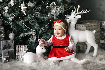A cheerful baby in a red outfit sits among Christmas decor under a festive tree, exuding joy and holiday spirit. HD wallpaper