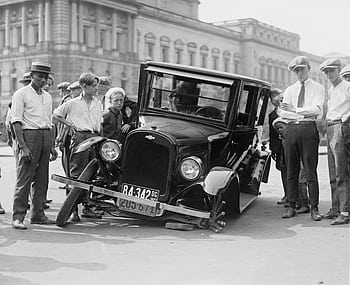 A vintage car crash attended by a group of onlookers in a 1920s urban setting. HD wallpaper