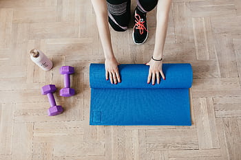 Top view of a woman rolling out a blue yoga mat with dumbbells and water bottle nearby for a home workout. HD wallpaper