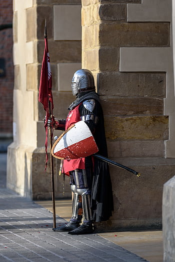 A medieval knight in armor stands guard with a shield and flag outside a stone building. HD phone wallpaper
