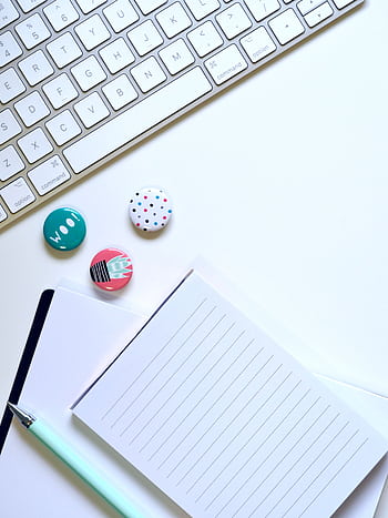 Flat lay of a minimalist workspace featuring a keyboard, colorful badges, and a notepad on a white background. HD phone wallpaper