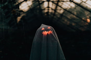 A ghostly figure with a glowing pumpkin face under a sheet in an eerie, dark setting. Perfect Halloween ry. HD wallpaper