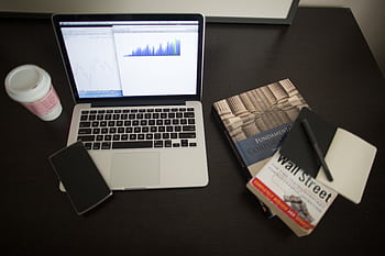Top view of a desk with laptop, charts, books, and coffee, depicting a modern financial workspace setup. HD wallpaper