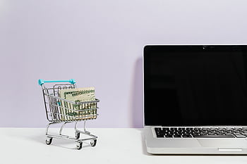 Mini shopping cart with cash beside a laptop, symbolizing online shopping and e-commerce. HD wallpaper