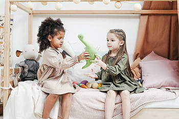 Two young girls sharing a plush dinosaur while sitting on a bed in a cozy playroom. HD wallpaper
