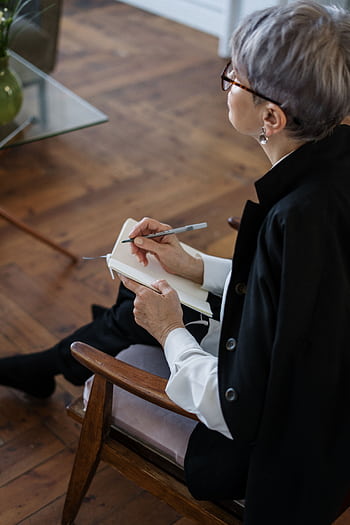 A professional woman with glasses taking notes while seated indoors, depicting consultation or therapy. HD phone wallpaper