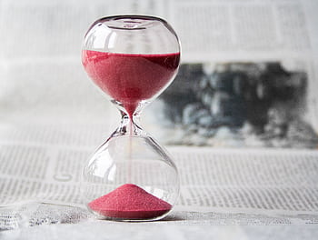 Close-up of a transparent hourglass with pink sand flowing, placed on a newspaper background. HD wallpaper