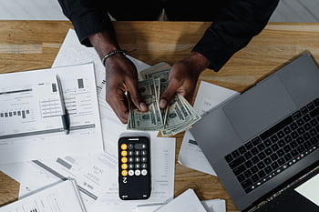 Person counting US dollars, using a calculator and laptop, with financial documents on a wooden desk. HD wallpaper