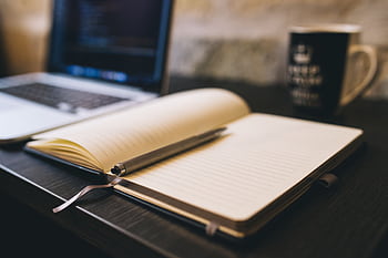 Open notebook with a pen on a wooden desk beside a laptop, ideal for blogging and work-related themes. HD wallpaper