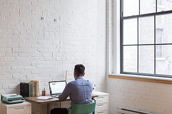 Adult male working remotely on a laptop in a well-lit home office with a large window. HD wallpaper