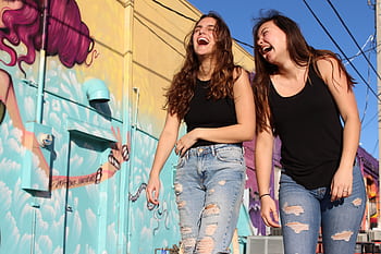 Two women laughing and enjoying a sunny day by a vibrant street mural. HD wallpaper