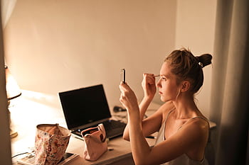 A young woman applies makeup at her vanity table, illuminated by soft light, in a cozy home setting. HD wallpaper