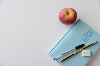 A minimalist flat lay of a study desk with a red apple, blue journal, pencil, pen, and eraser. HD wallpaper