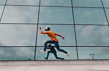 A man in motion jumps against a glass facade, creating a dynamic urban reflection. HD wallpaper