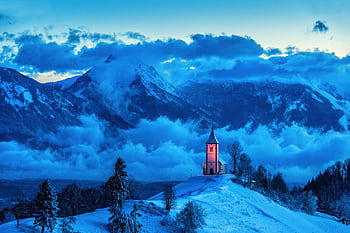 A serene winter scene of a church on a snowy hill with mountains and clouds during blue hour. HD wallpaper