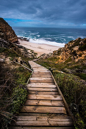 Wooden stairs leading down to a scenic beach surrounded by cliffs under a dramatic sky. HD phone wallpaper