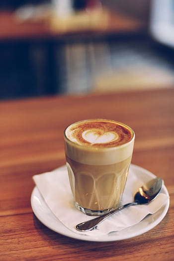 A close-up of a beautifully crafted latte in a glass cup, showcasing intricate foam art, on a wooden cafe table. HD phone wallpaper