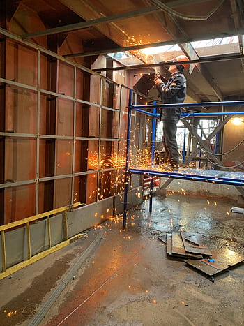 A construction worker welds metal indoors, creating flying sparks in a factory setting. HD phone wallpaper
