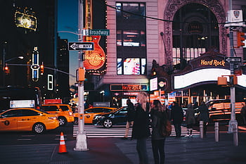A bustling street view of Broadway at night featuring Hard Rock Cafe, taxis, and pedestrians in New York City. HD wallpaper