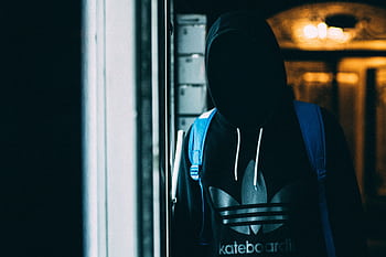 A shadowy figure in a hoodie stands with a backpack in dim lighting, conveying mystery and anonymity. HD wallpaper