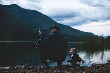 Father and children enjoy a serene moment by a lakeside with mountains. HD wallpaper