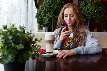 A young woman enjoys a latte while browsing her smart in a cozy cafe. HD wallpaper
