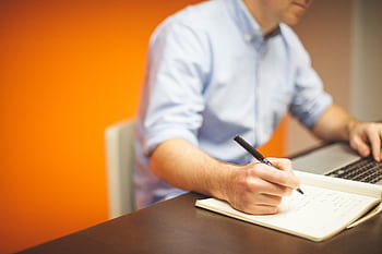 A man sits in modern office, writing notes with a laptop open. HD wallpaper
